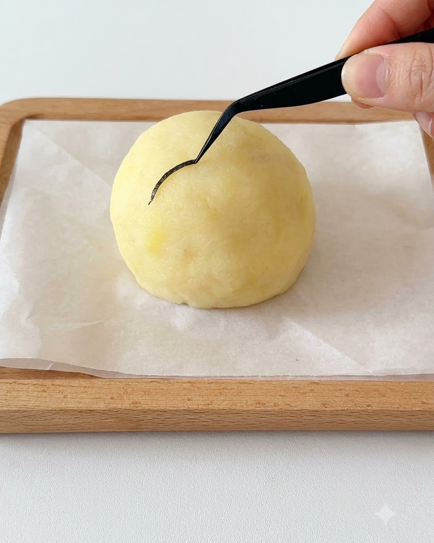 細いピンセットを持った手が、丸いポテトの頭に細くカーブしたのりを配置している様子。