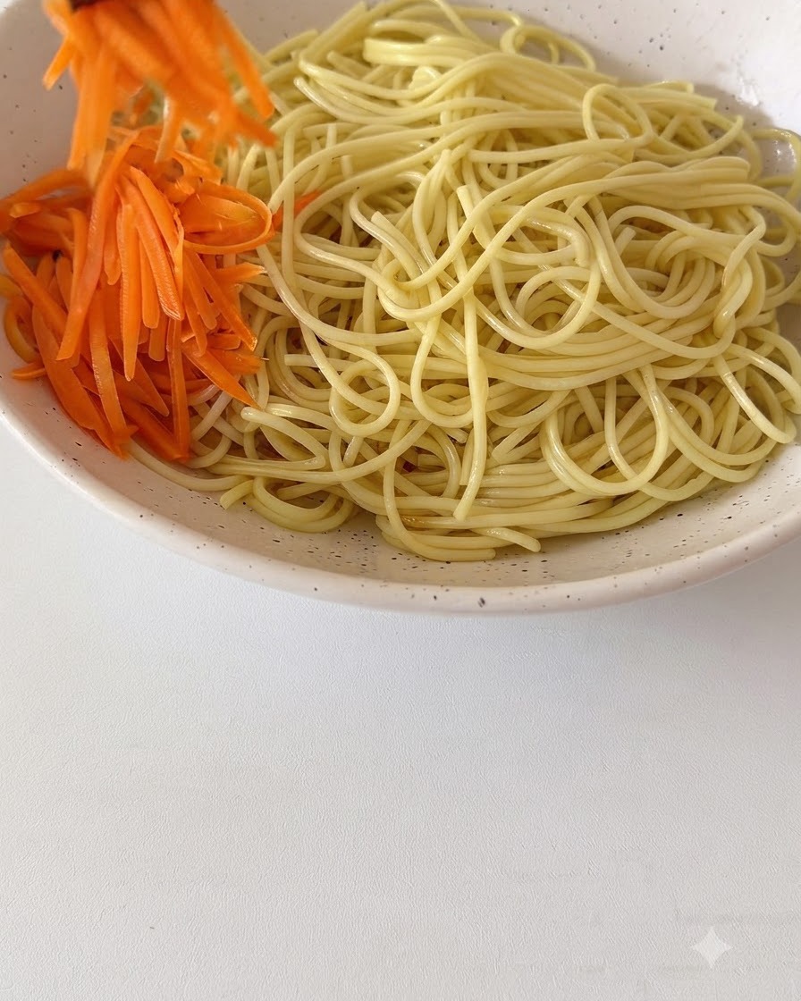 大きな陶器のボウルに入った茹でた麺の上に、新鮮な千切りにんじんや他のトッピングが並べられている様子。