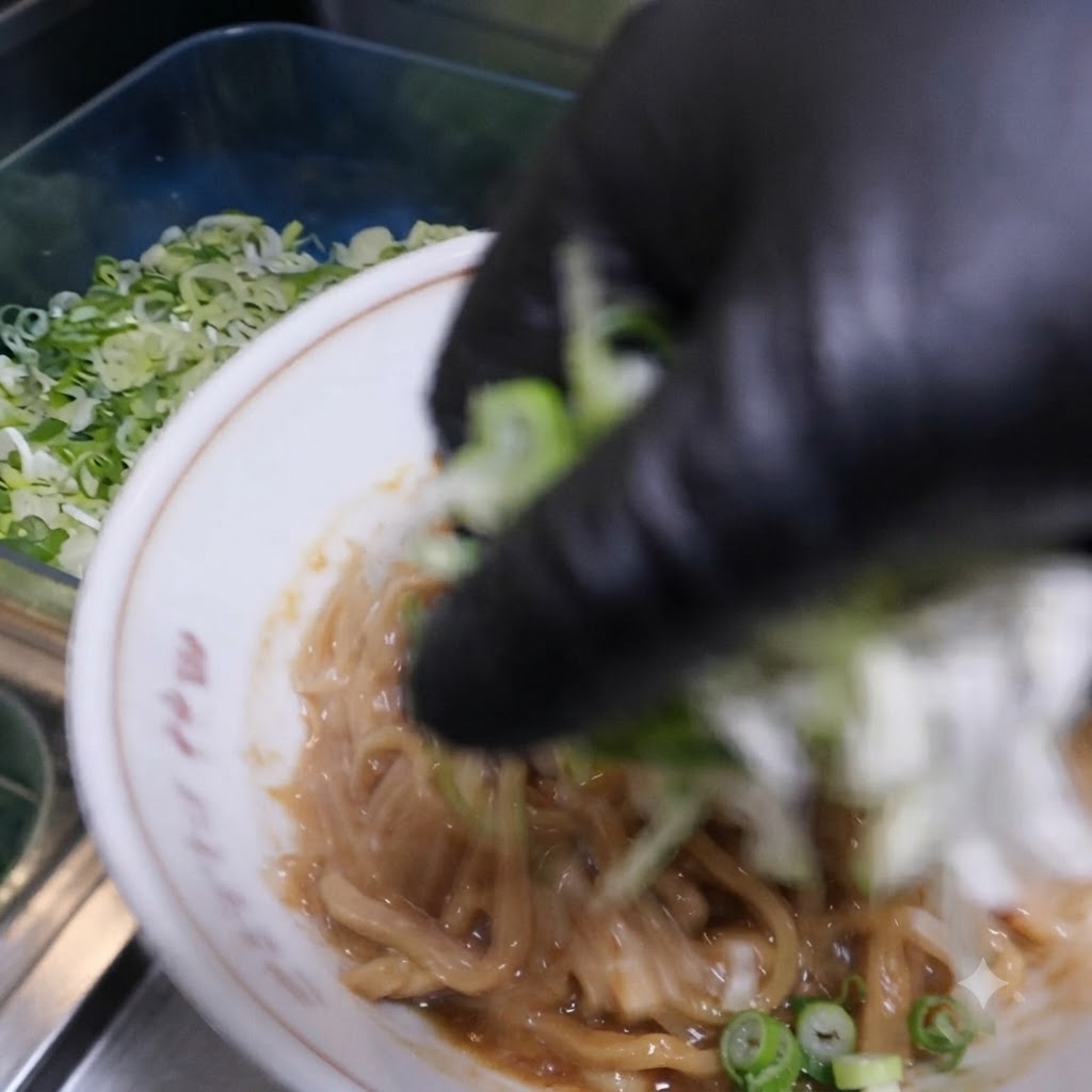 手袋をした手が、丼の中の味付けされた麺の上に刻んだ青ネギとニラを散らしている様子。