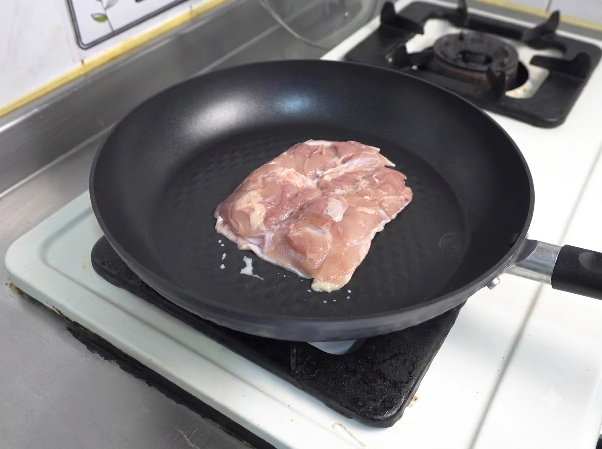 暗い色のノンスティックフライパンの皮を下にして生のエスニックな鶏もも肉が置かれている様子。