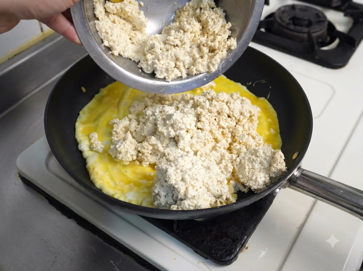 黒いフライパンの中で調理中の黄色い炒り卵の上に、金属製ボウルから崩した白い豆腐が入れられている様子。