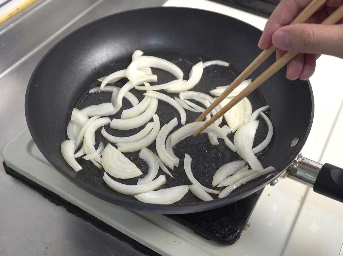 木製の箸を使ってフライパンでスライスされた白玉ねぎが炒められている様子。
