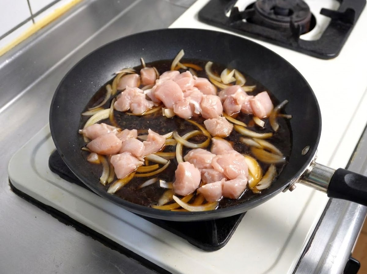 玉ねぎと一緒に煮立っている濃い色の煮汁の中に生の鶏肉のピースが入れられている様子。