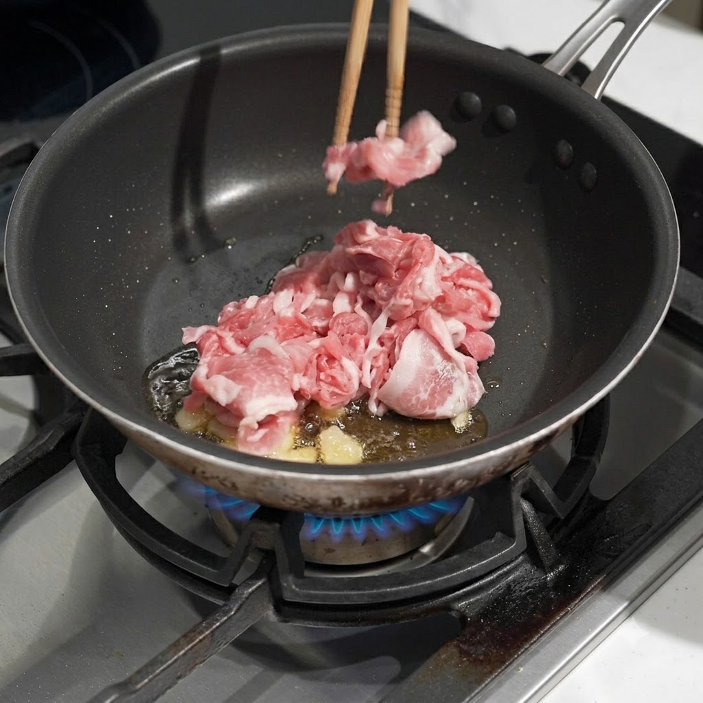 熱した油とにんにくが入ったフライパンに、生の豚肉を入れ、菜箸で炒めている様子