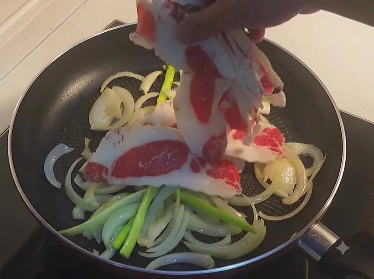 スライスした玉ねぎと青ねぎが入ったフライパンに、生の牛バラ肉の薄切りを入れている様子。