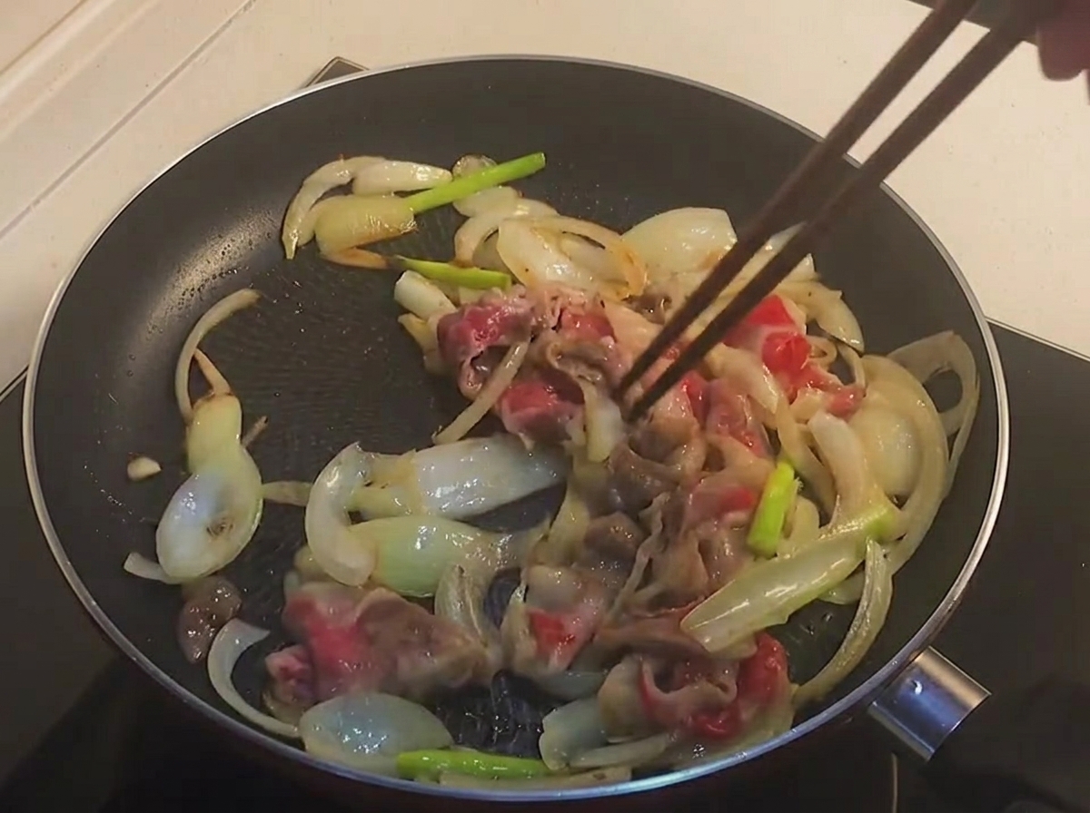 黒いフライパンの中で、軽く焼き色がついた半生状態の牛バラ肉と玉ねぎを箸で炒めている様子。