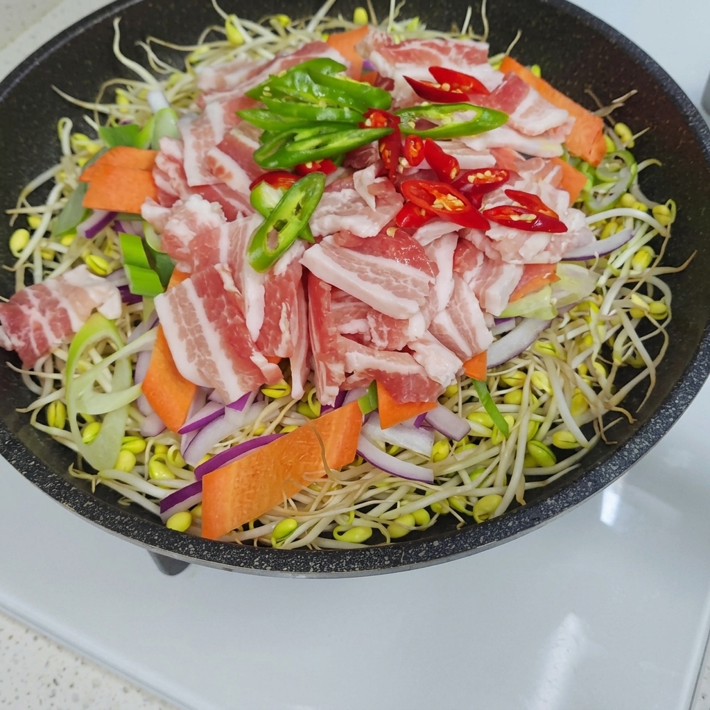 もやしを敷き、その上にスライスした玉ねぎ、にんじん、生の豚バラ肉、緑と赤の唐辛子を重ねたフライパンの俯瞰図。