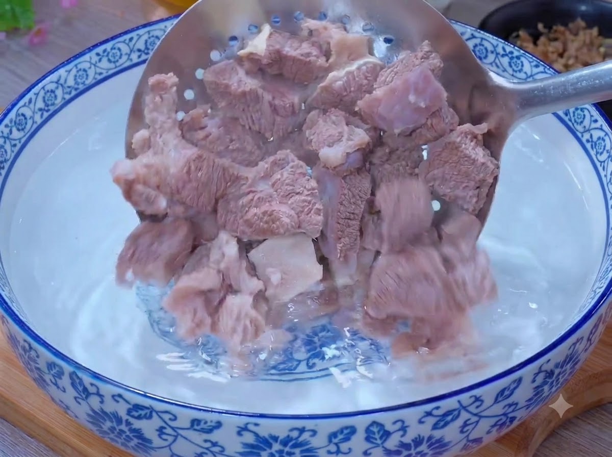 ボウルに入ったぬるま湯の中で、穴あきお玉を使って下茹でされた牛肉の破片を洗っている様子。