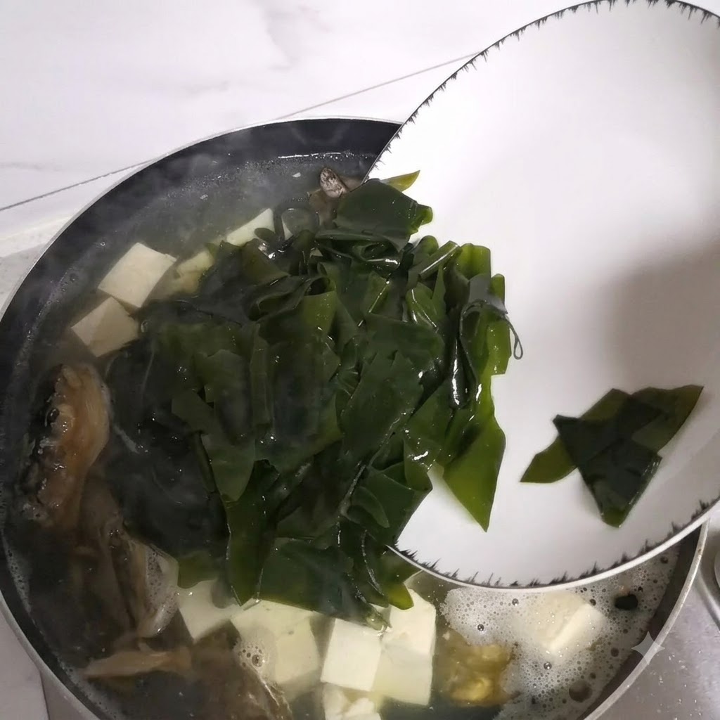 白い皿から、魚の頭と豆腐が入った煮込み中の鍋に海藻が滑り落ちている様子。