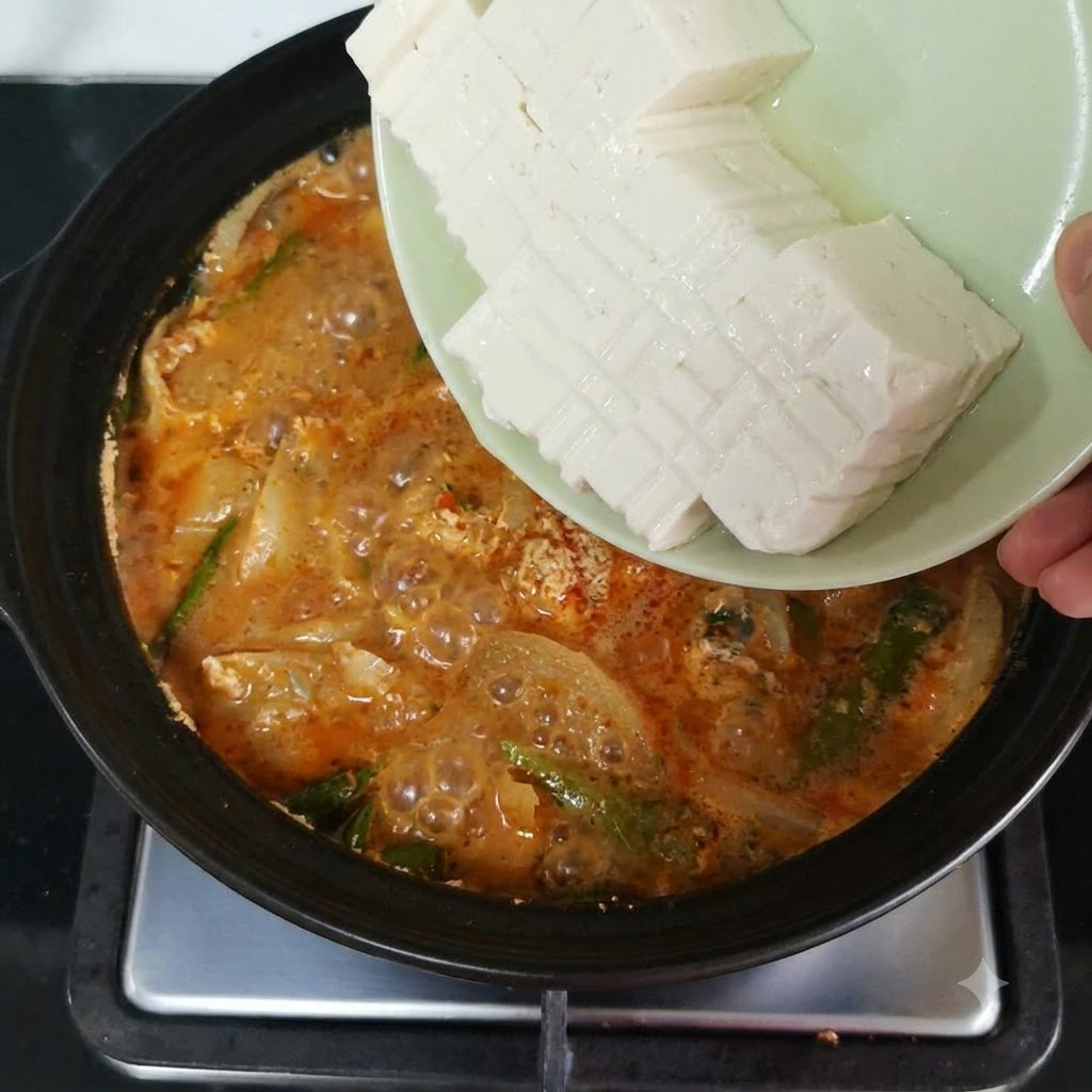 オレンジ色のピリ辛スープと野菜が入った鍋に、スライスした絹豆腐を皿から滑り込ませている様子。