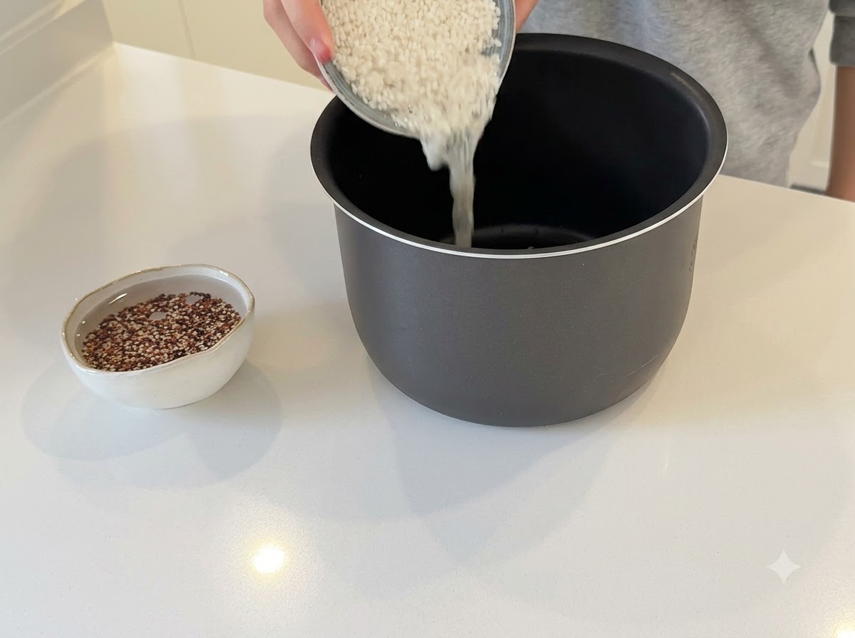 黒い炊飯器の内釜に白米を注いでいる手と、水に浸したキヌアの小さなボウル。