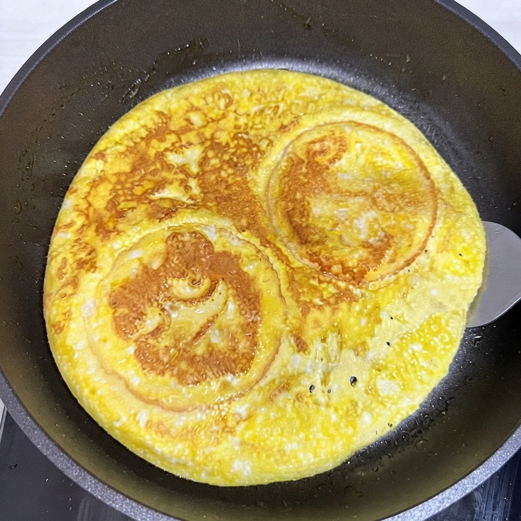 フライパンの中で、スパチュラを使って丸く焼き上がった卵とバンズの層を裏返しているところ。
