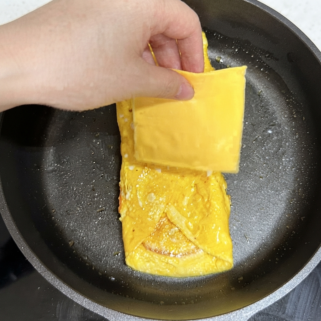 フライパンの中の焼き上がった卵の上に、四角いスライスチーズを置いている手。