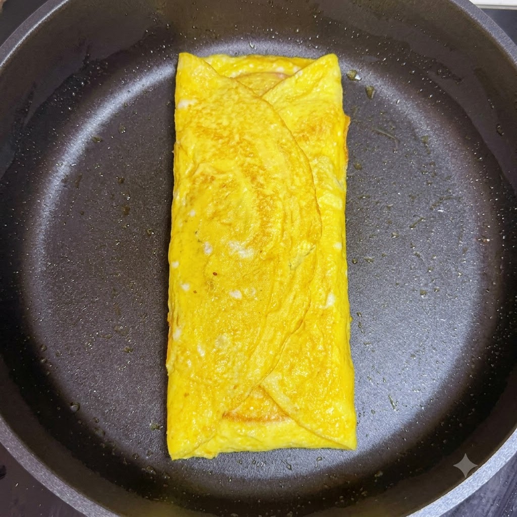 フライパンの中の卵に包まれたチーズの上に、折りたたんだピンク色のハムを重ねているところ。