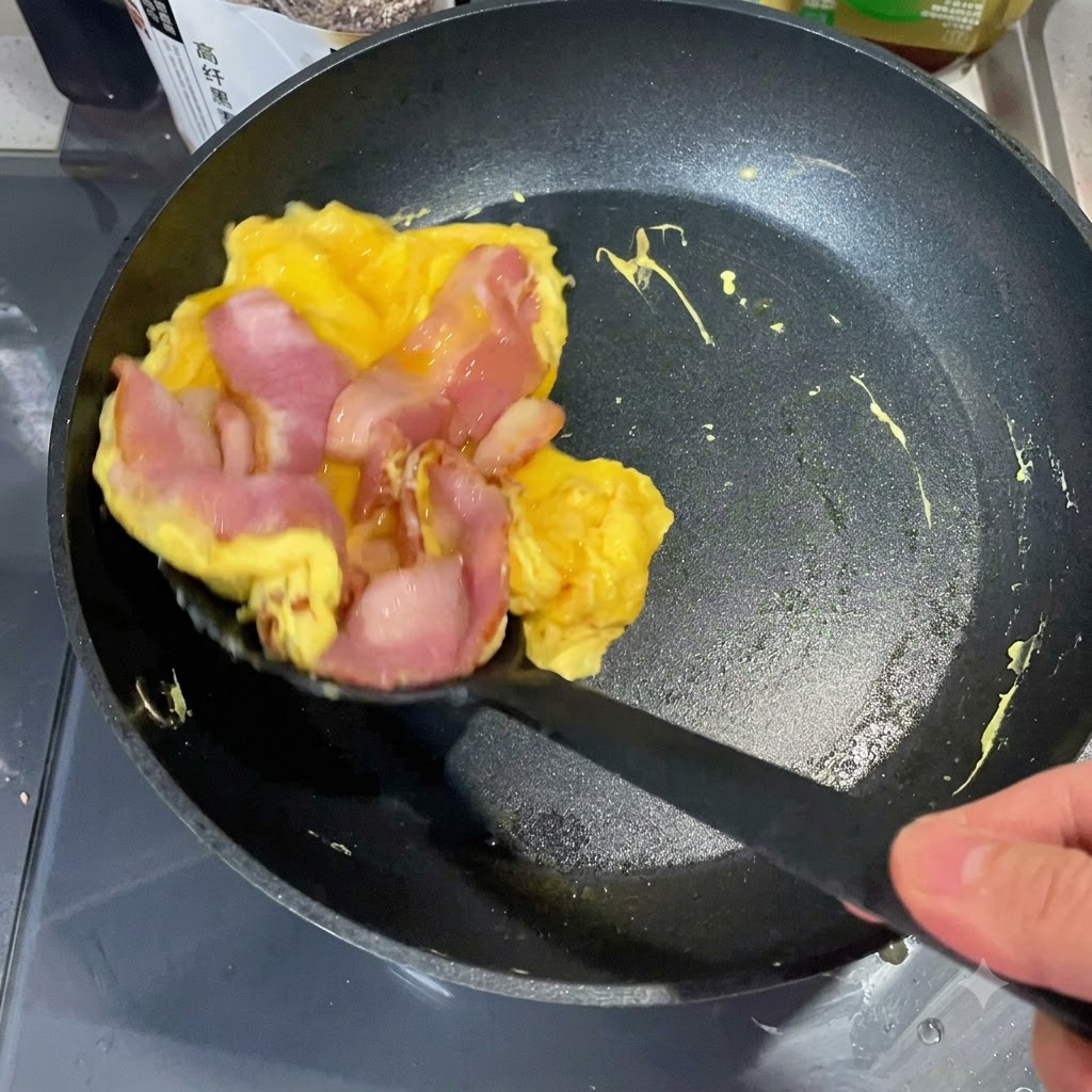 スパチュラが調理した卵とベーコンのスタックを持ち上げ、トーストしたイングリッシュマフィンの半分の上に載せようとしている様子。