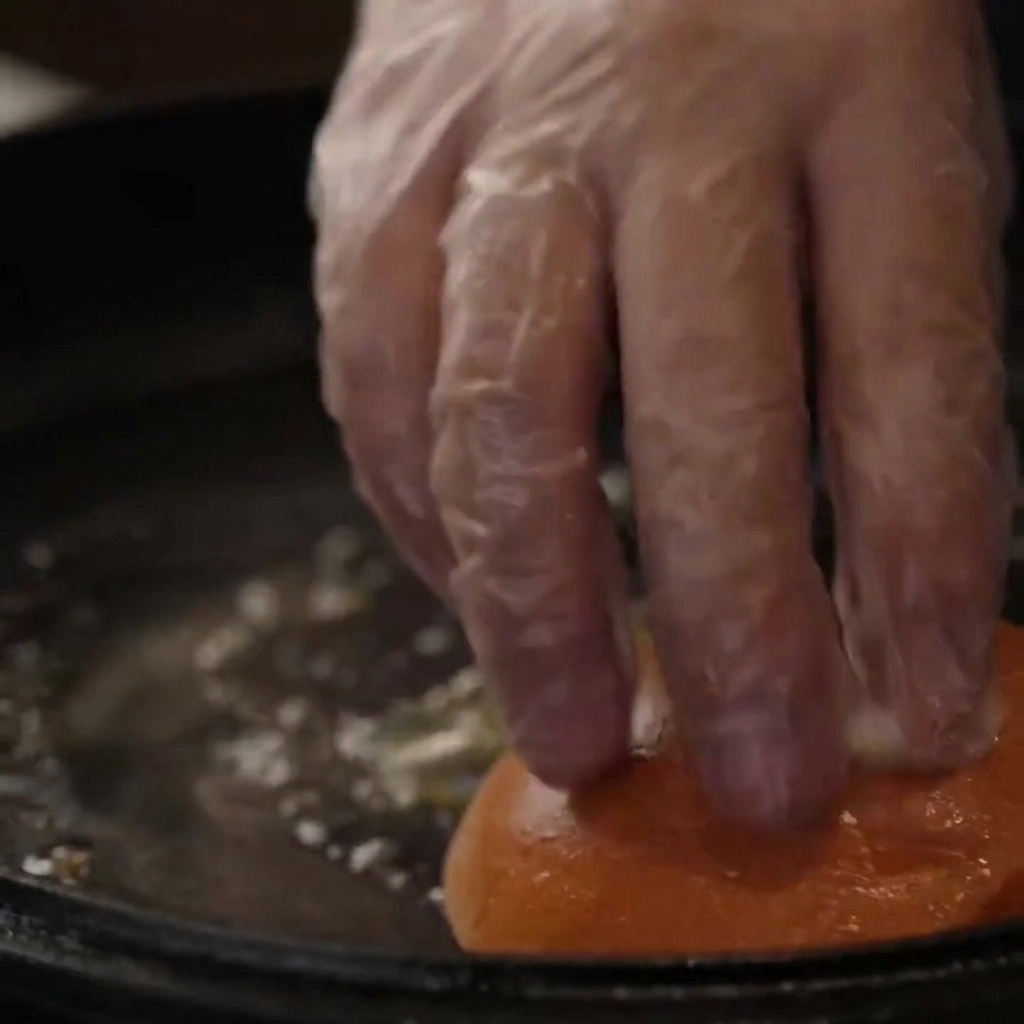 手袋をした手が、トーストのためにバターを塗った鋳鉄製スキレットにバーガーバンズを押し込んでいるところ。