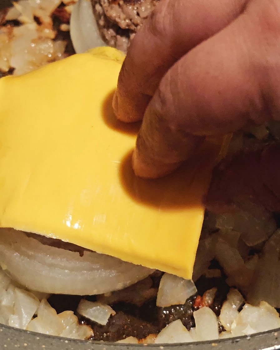 フライパンで調理された肉詰めオニオンリングの上に、黄色いスライスチーズを乗せている様子。