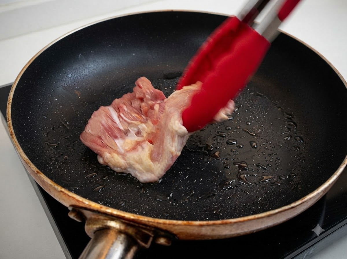 赤いトングを使って、油を引いた熱したフライパンに生の鶏もも肉を投入している様子。