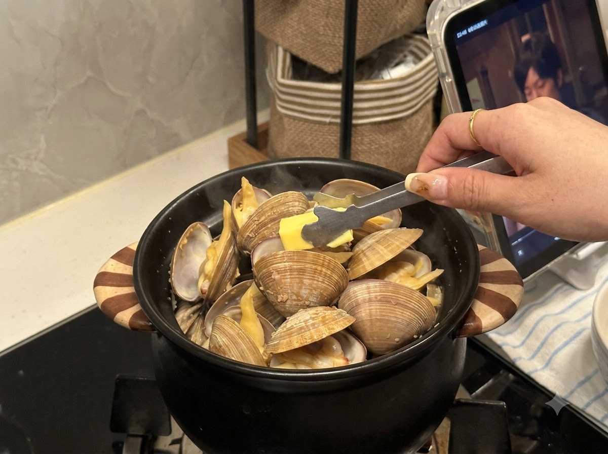 開いたあさりの詰まった黒い鍋の中に、トングで小さめのバターを投入している様子。