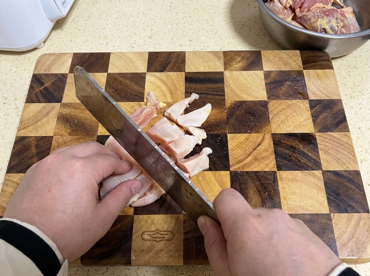 格子模様の木製まな板の上で、包丁を使って生の鶏胸肉を小さく切っている様子。