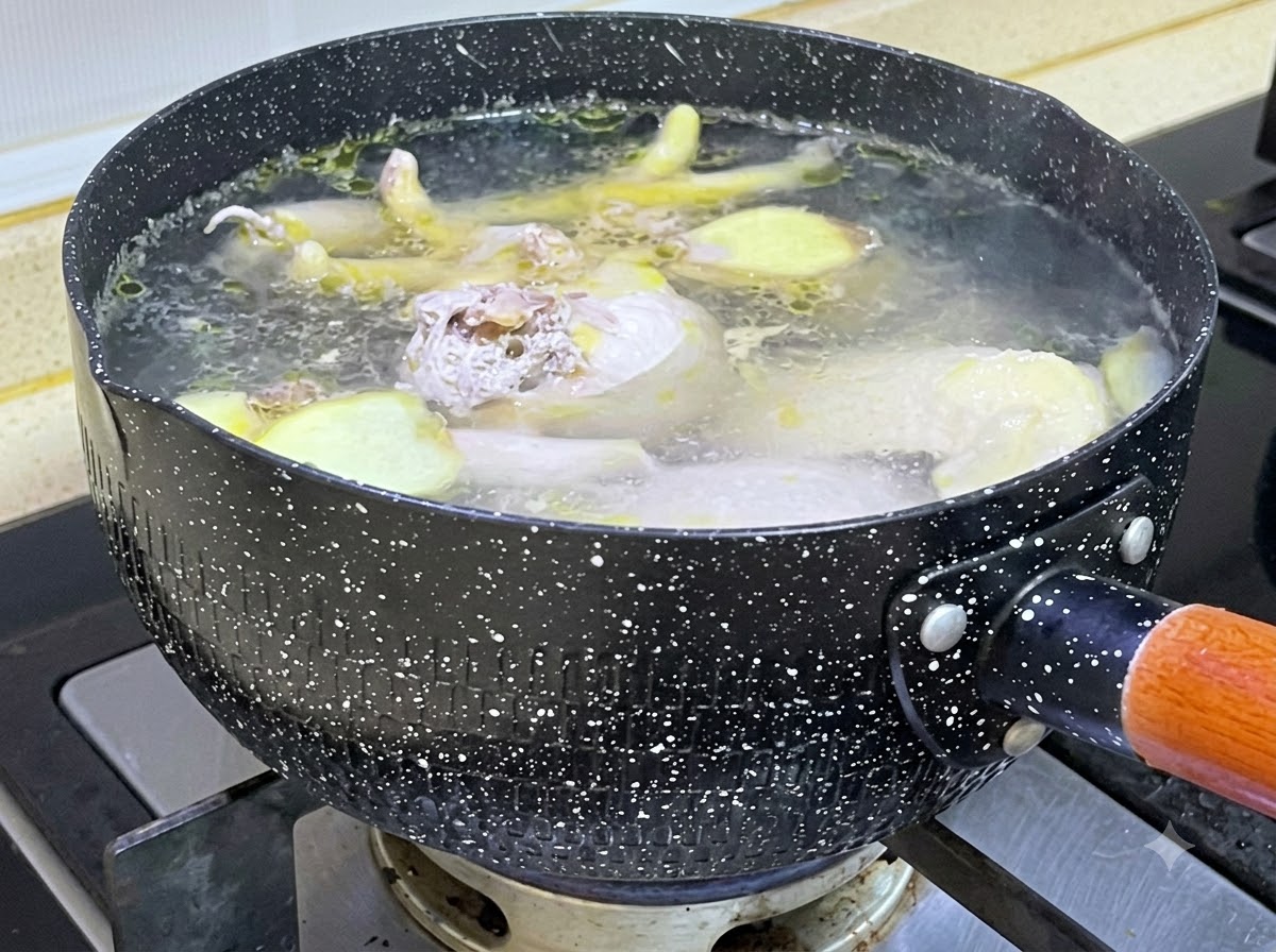 沸騰したお湯が入った黒い speckled ポットの中で、生の鶏肉と生姜のスライスが煮込まれている様子。