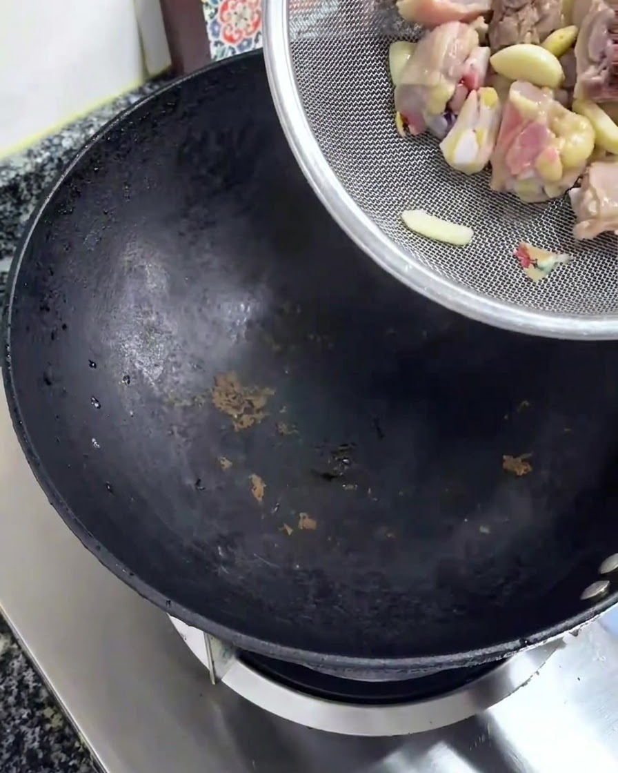銀色のザルに入った生のぶつ切り鶏肉が、黒い中華鍋に注がれている様子。