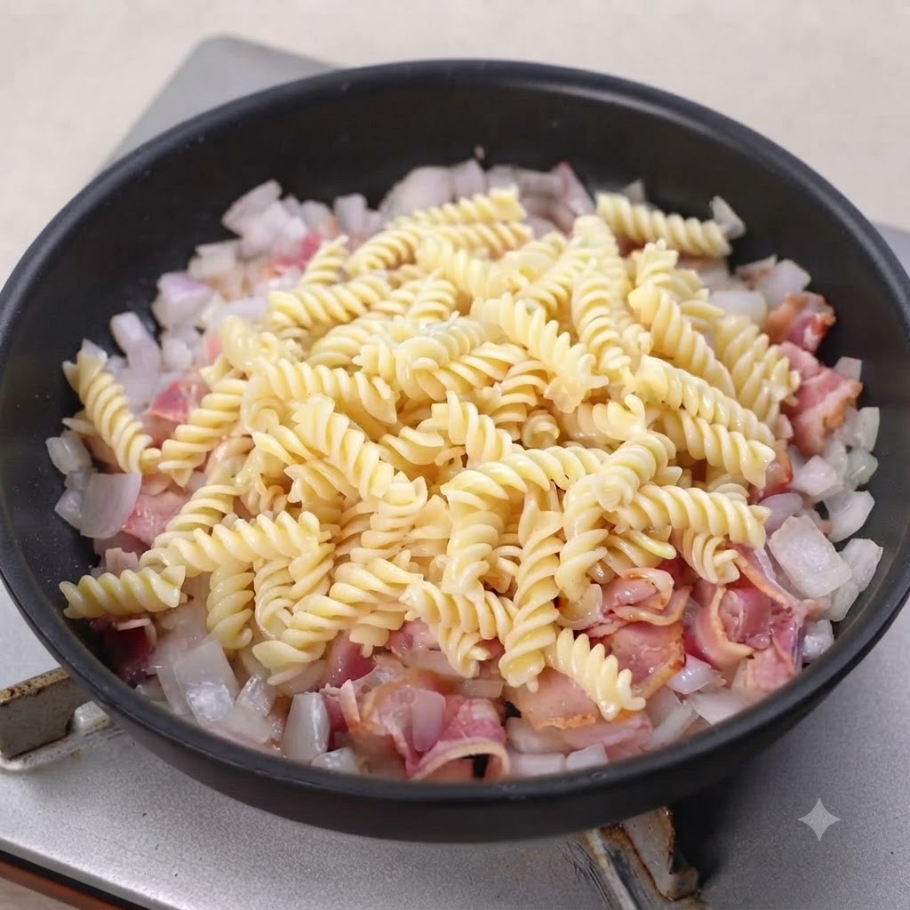 炒めたベーコンと玉ねぎの上に茹でたショートパスタを乗せている黒いフライパン。