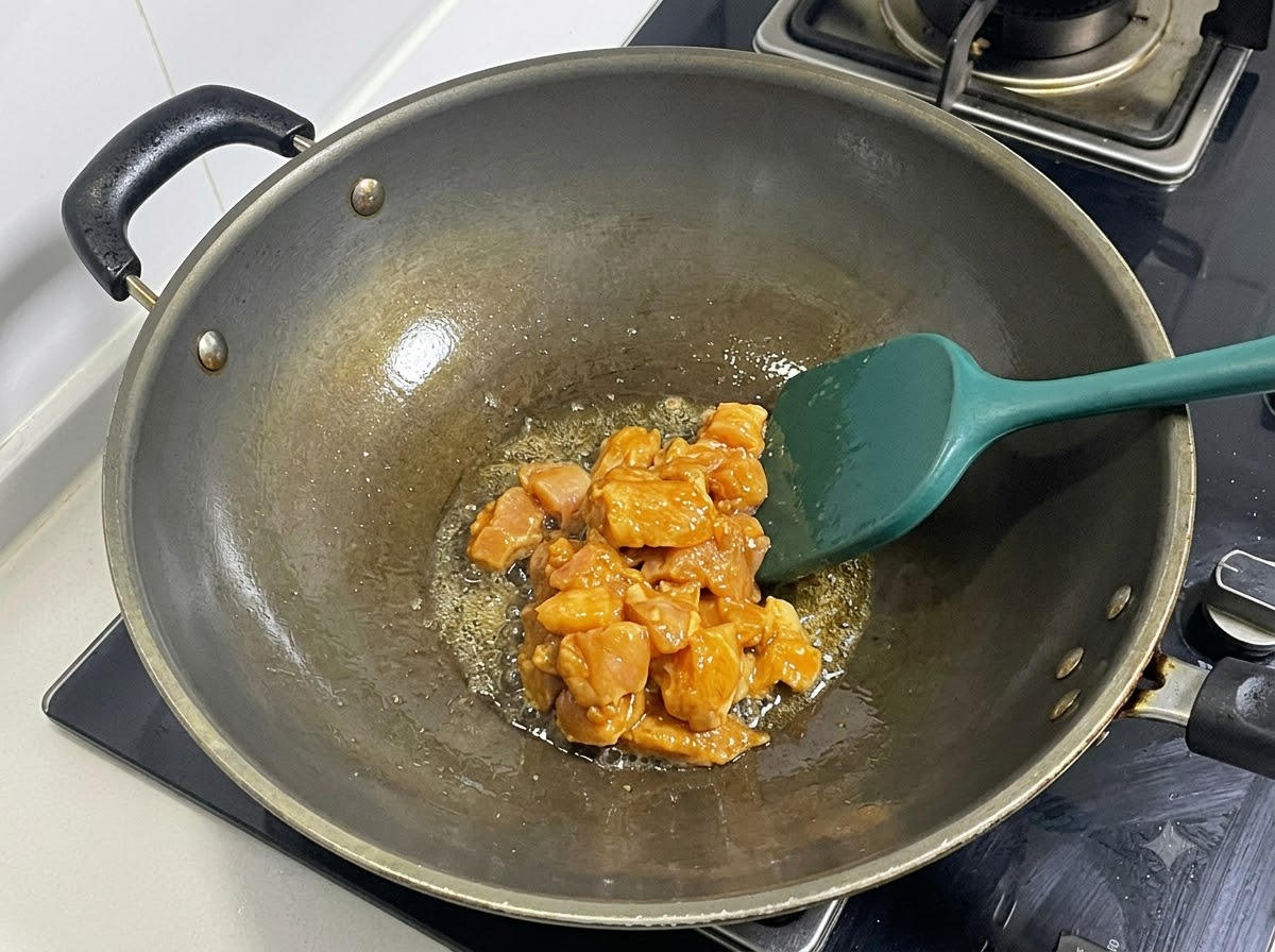 緑色のヘラを使い、熱した中華鍋で下味をつけたサイコロ状の鶏むね肉を炒めている様子。