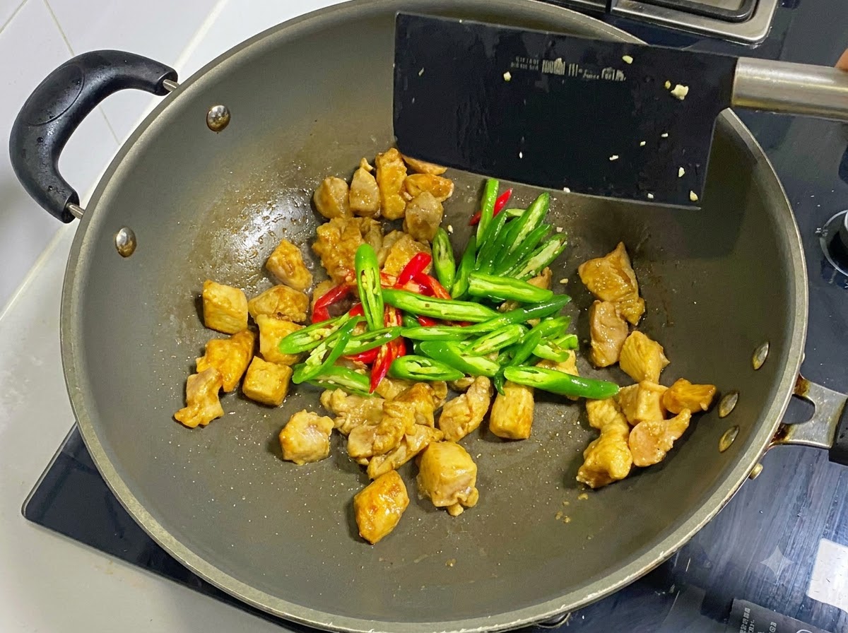 黒いヘラを使い、中華鍋で炒めた鶏肉にスライスした青唐辛子と赤唐辛子を加えて混ぜ合わせている様子。