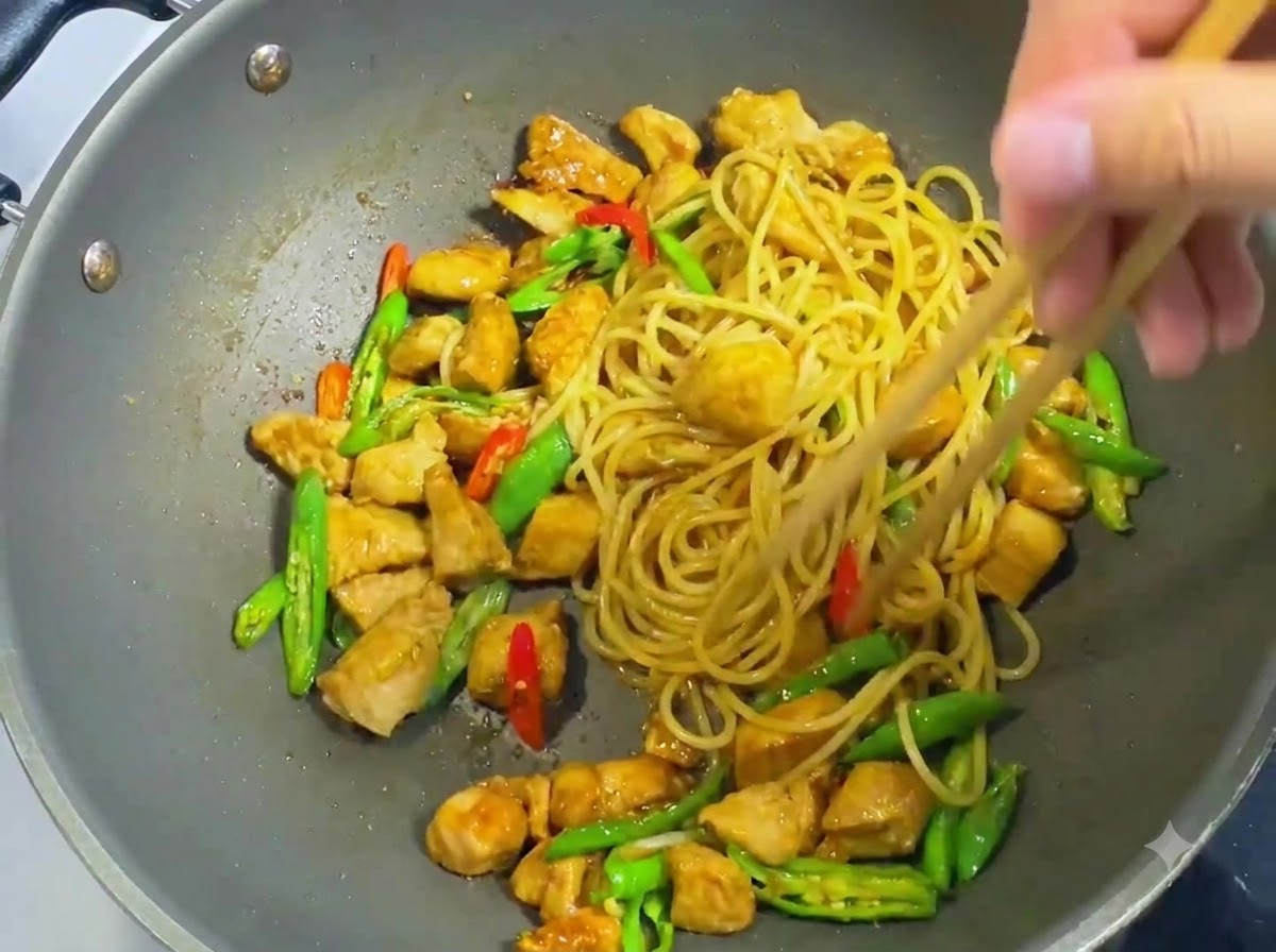 木製の菜箸を使い、中華鍋の中でスパゲッティ、鶏肉、唐辛子、濃い色のソースを均等に和えている様子。