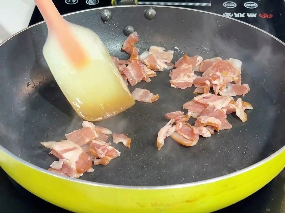 フライパンの中で、スパチュラを使ってベーコンと玉ねぎを炒めている様子。