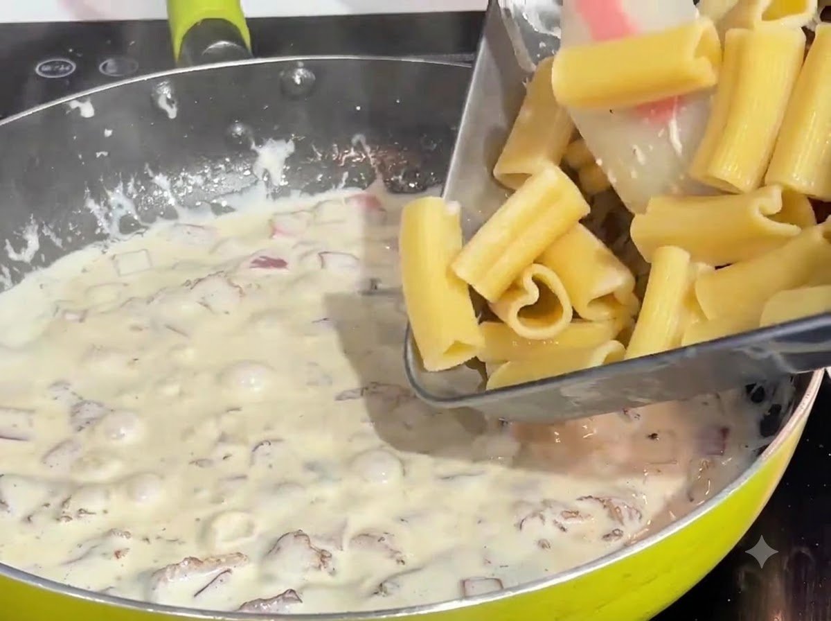 トレイに入った茹で上がったハート型パスタを、フライパンの沸騰したクリームとベーコンのソースに投入している様子。