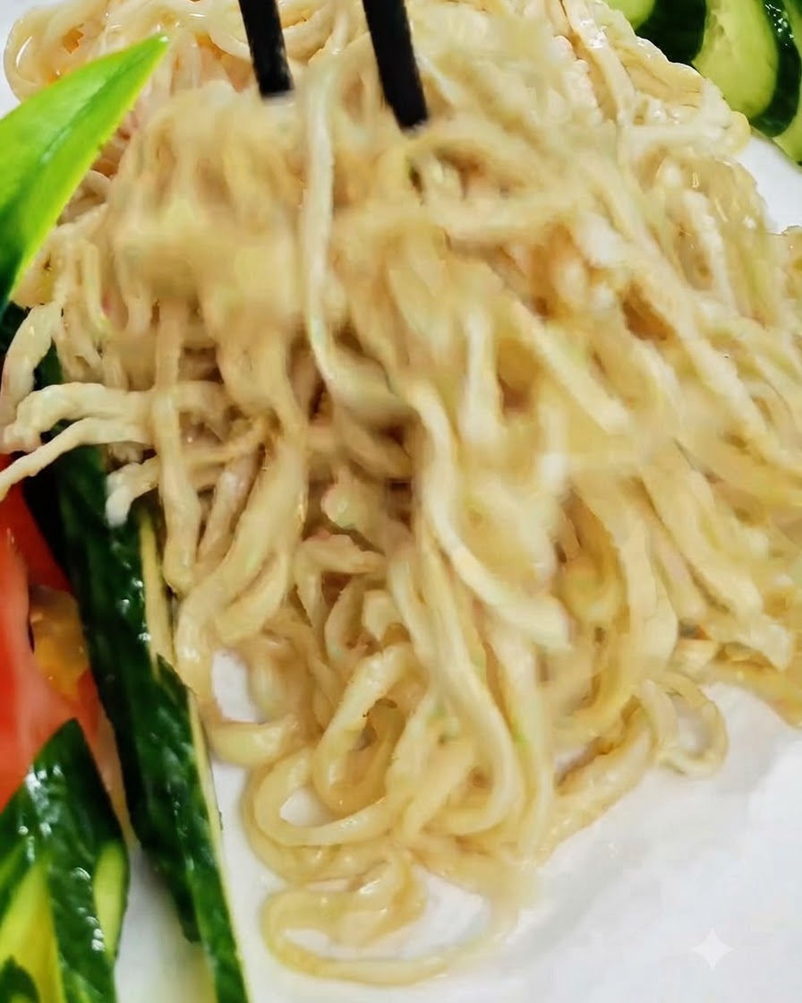 黒い箸を使って、きゅうりで飾られた白い皿に調理済みの麺を持ち上げて盛り付けている様子。