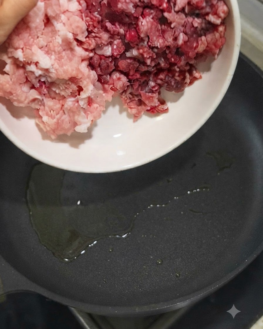 白いボウルから油を熱したスキレットに豚ひき肉と牛ひき肉を投入しているところ。