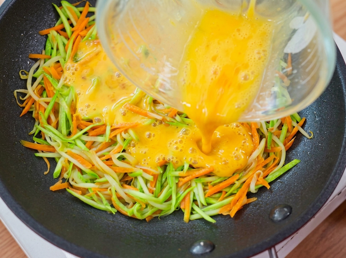 フライパンに敷いた野菜の上に計量カップから溶き卵を注ぎ入れている様子。