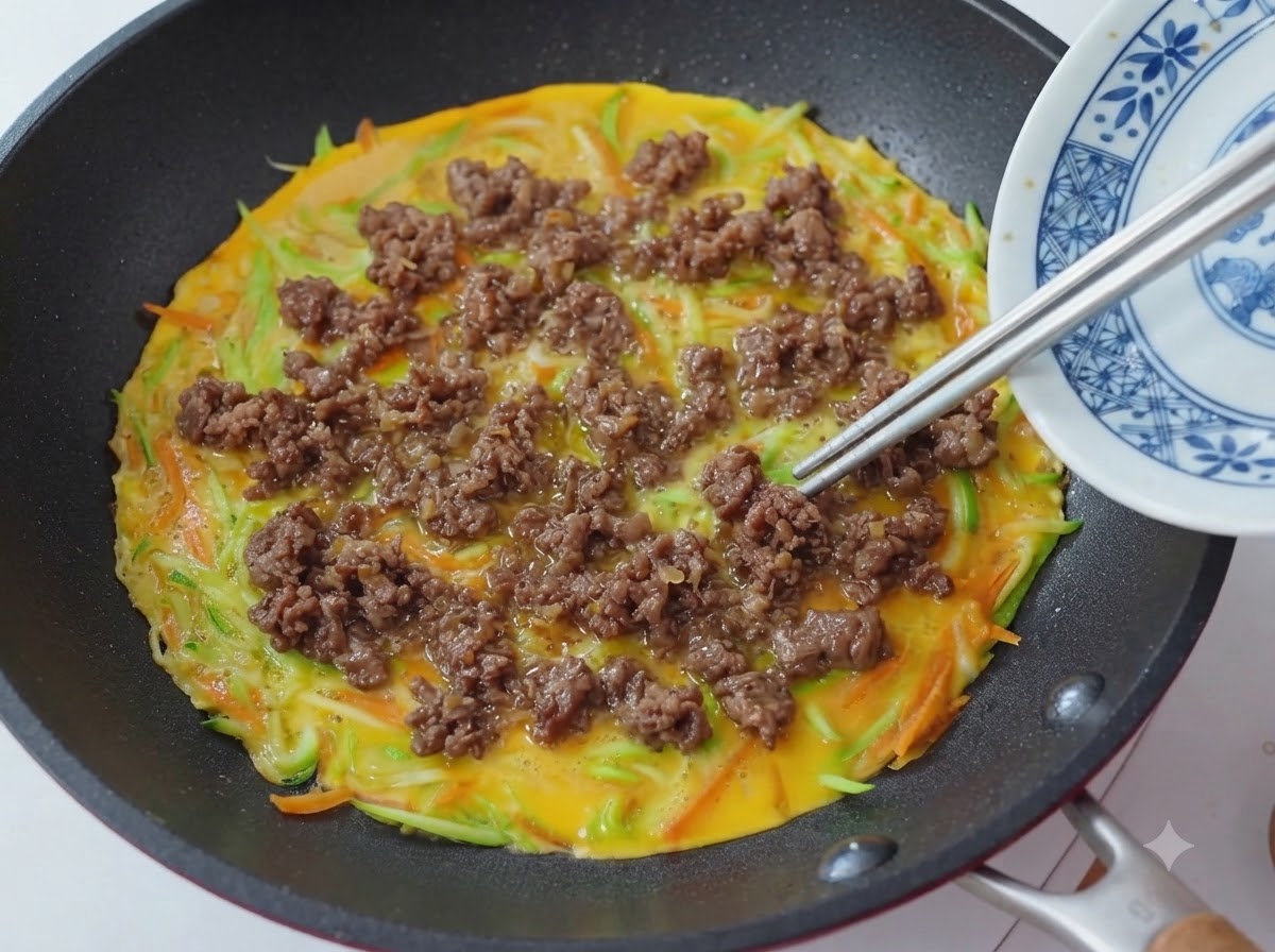 野菜と卵の生地の上に箸で炒めた牛肉を均等に散らしてのせている様子。