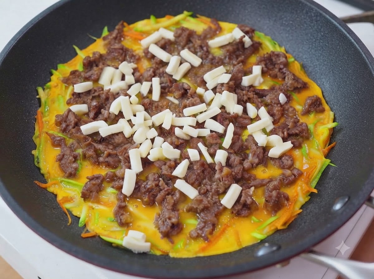 フライパンの牛肉と卵の上に細切りのモッツァレラチーズを振りかけている様子。