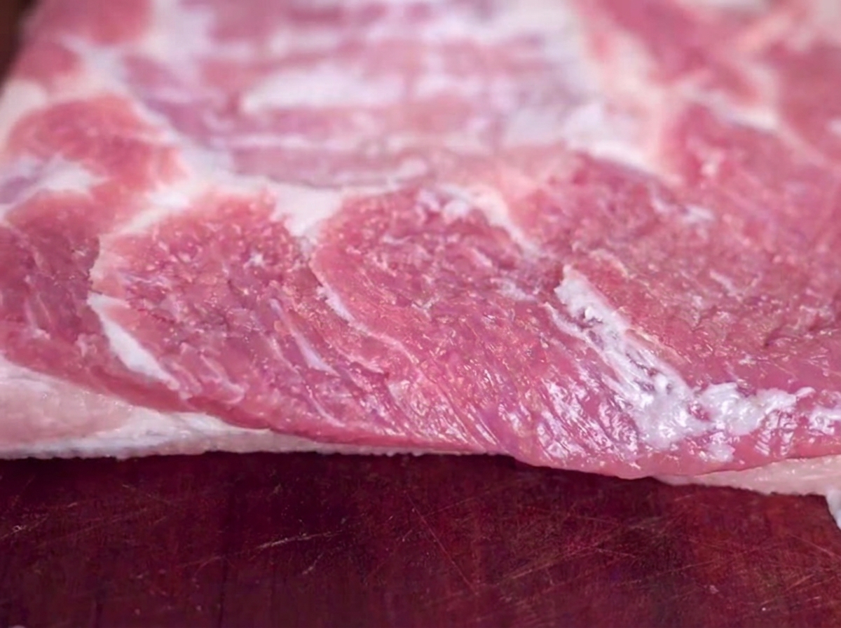 木のまな板の上に置かれた豚バラ肉ブロックに塩を振っている様子