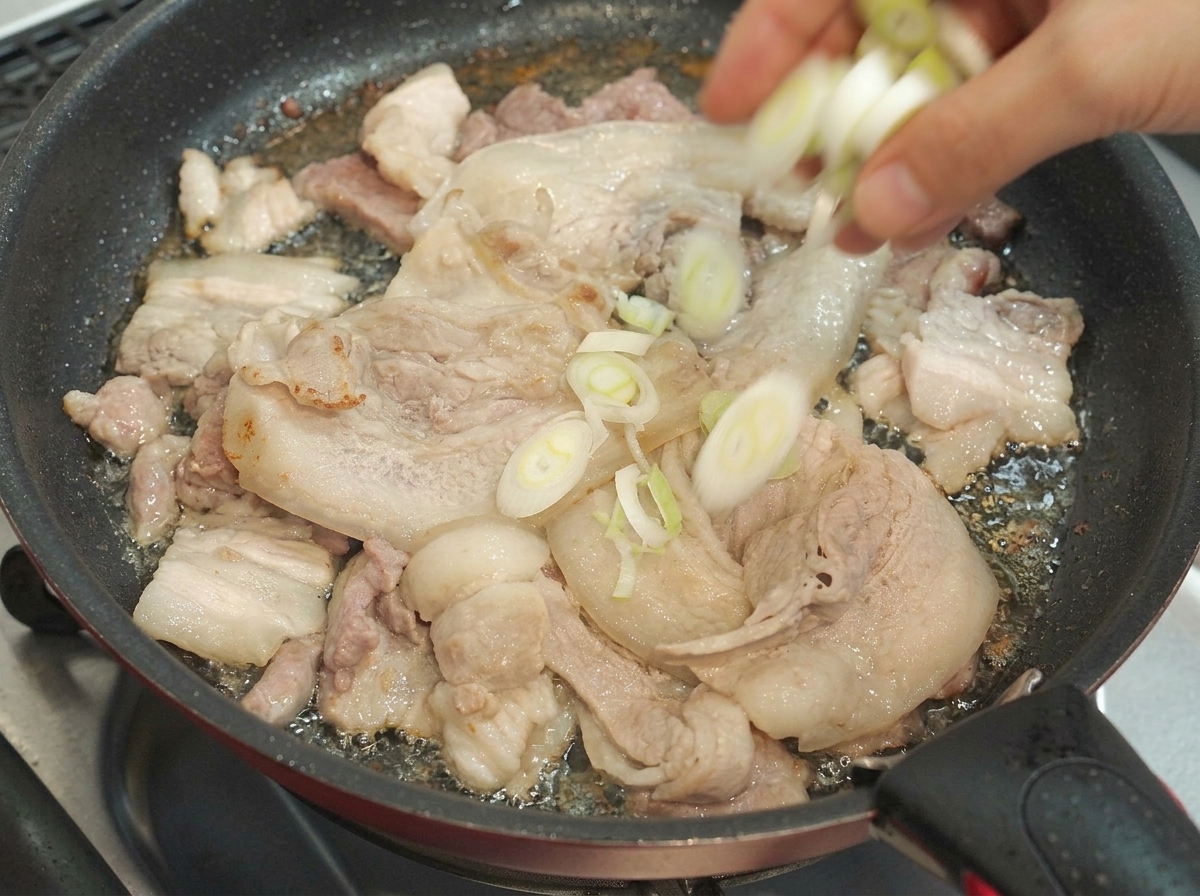 調理中の豚バラ肉のスライスの入ったフライパンに刻んだ青ネギが入れられている様子