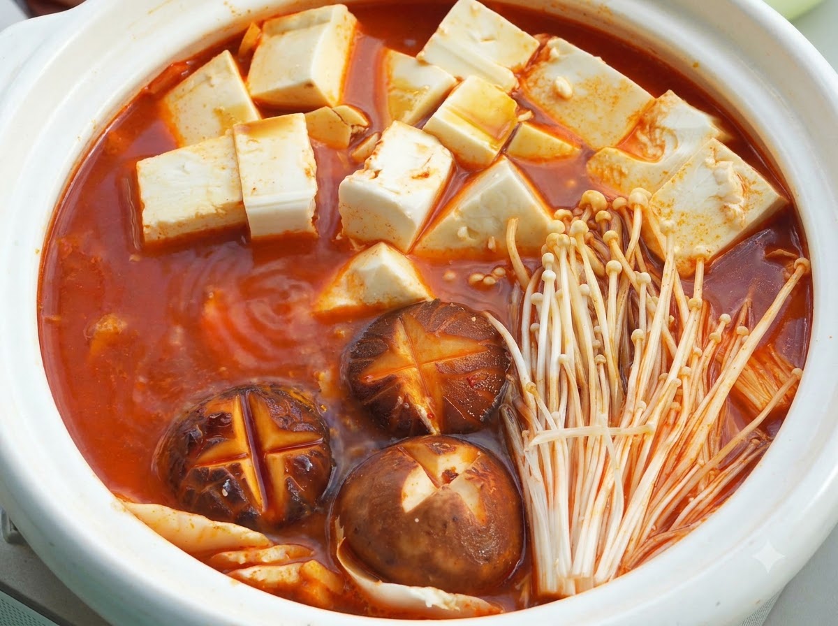 蓋の下で、赤いスープベース、絹ごし豆腐のキューブ、十字の切り込みを入れたシイタケ、エノキタケが入った白いセラミック製の鍋。