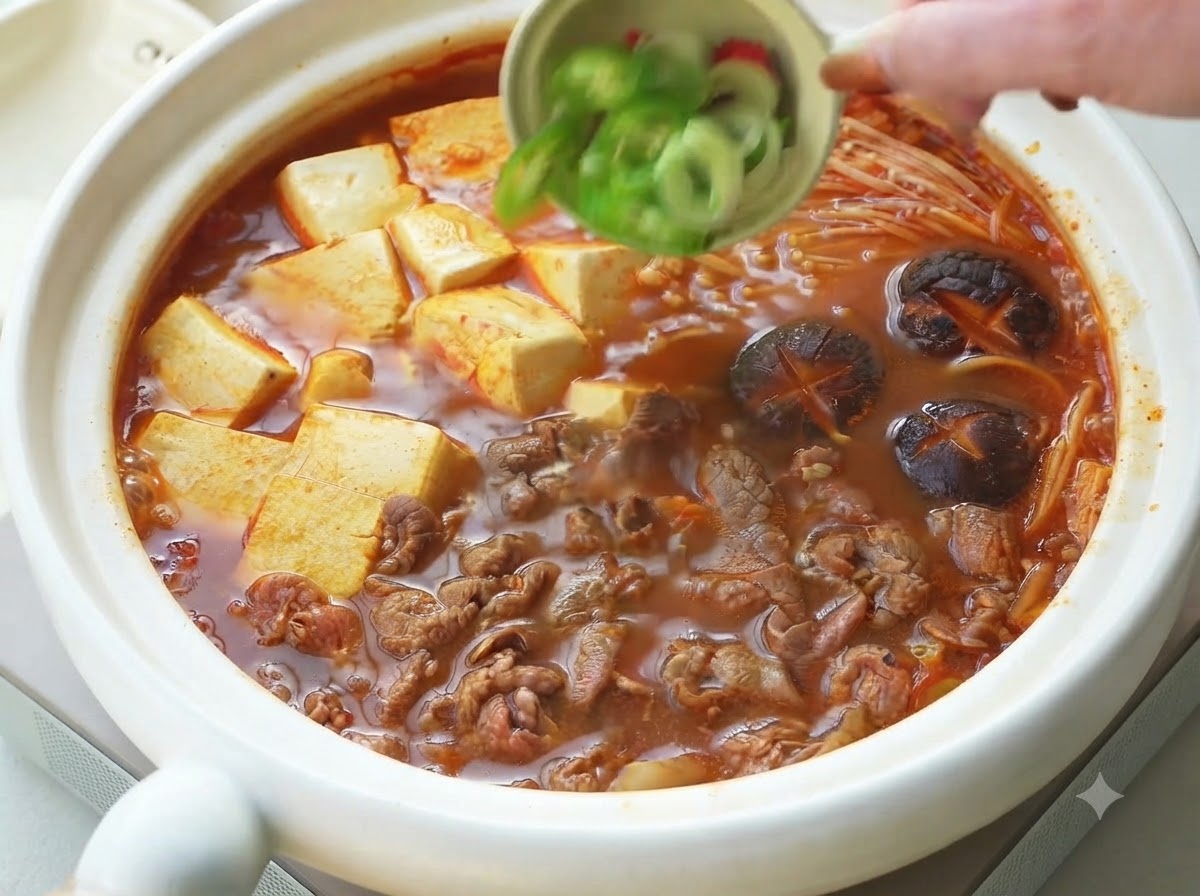 調理された韓国風牛肉豆腐チゲの上に、小さなボウルから刻みネギとスライスした唐辛子をトッピングとしてかけている様子。