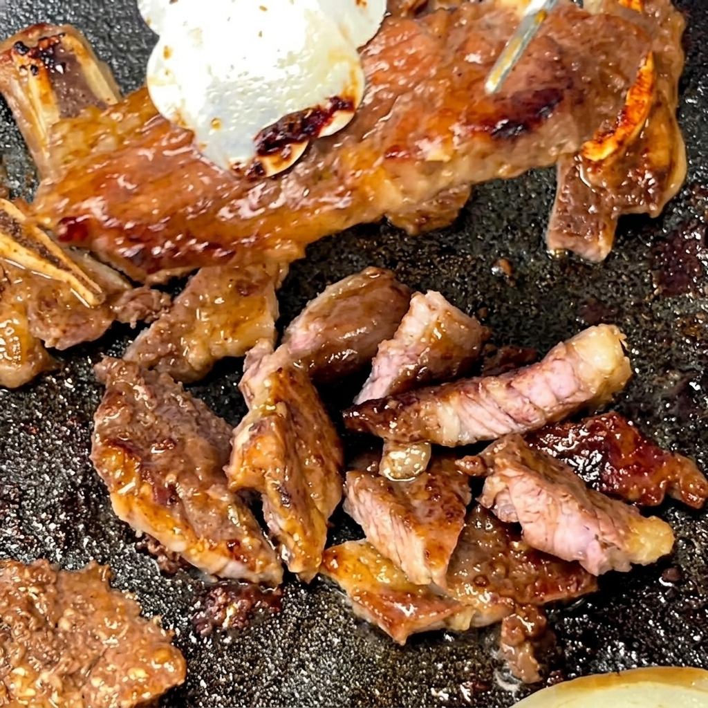 トングで焼き上がったショートリブをつまみながら、熱いフライパンの上でキッチンバサミを使って一口大に切り分けている様子。