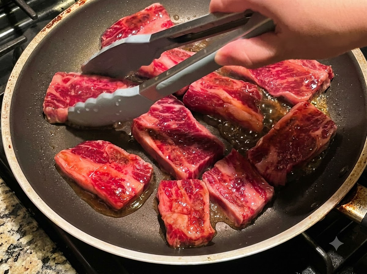 フライパンで焼けている複数の牛カルビをトングで裏返している様子。