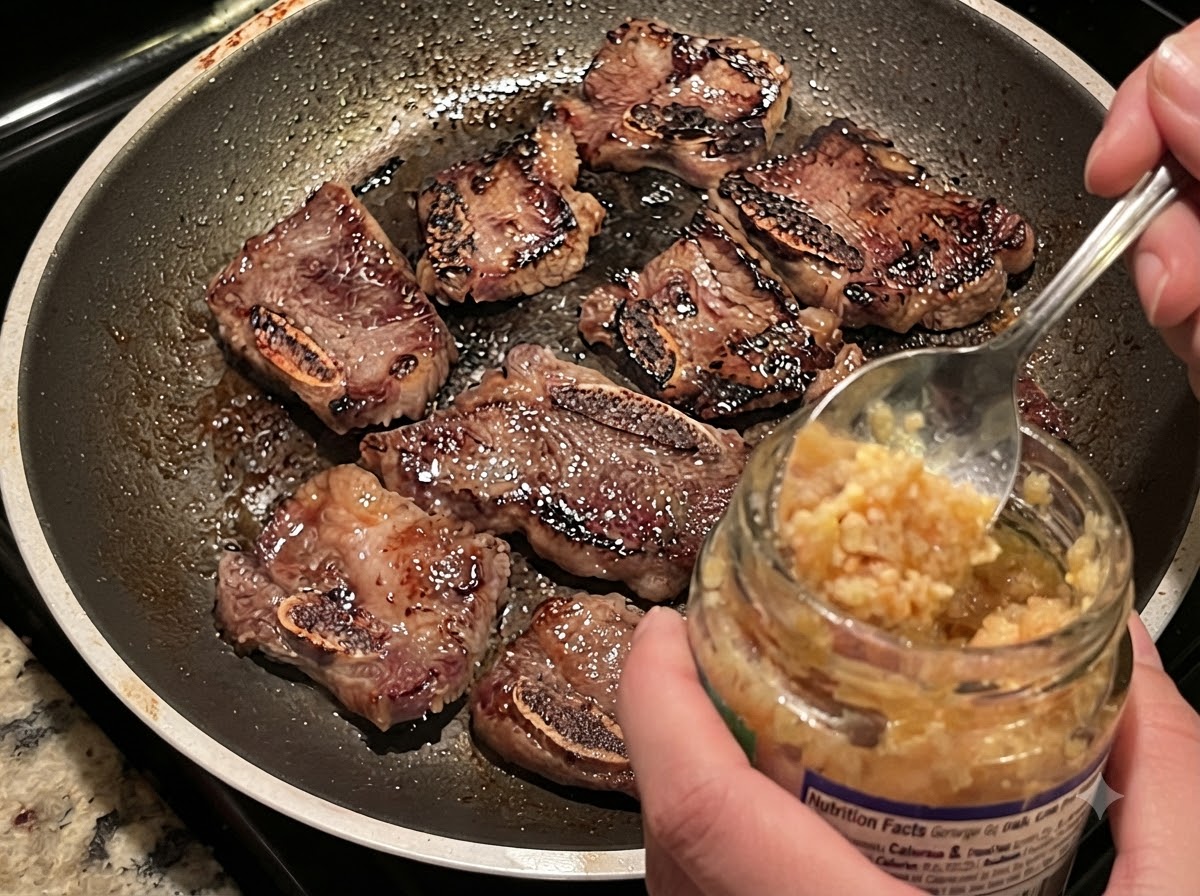 焼き色のついた牛カルビと玉ねぎが入った熱々のフライパンに、瓶からスプーンでにんにくのみじん切りを加えている様子。