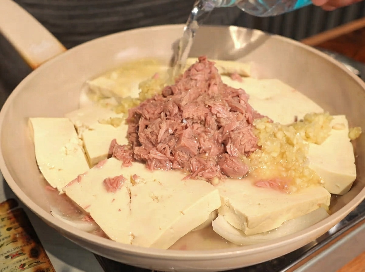 ボトルから透明な水が、豆腐、玉ねぎ、ニンニク、ツナの入った鍋に注がれている様子。