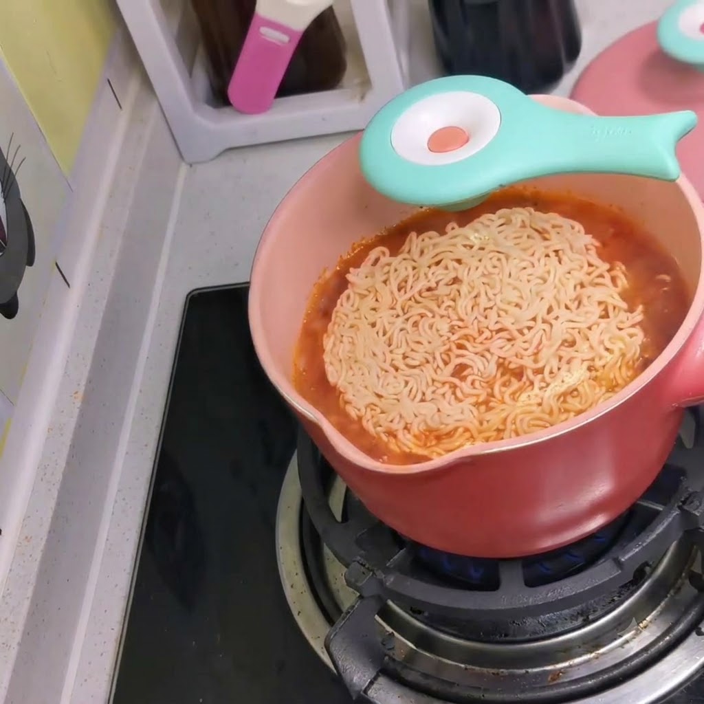 コンロ上の沸騰した赤い辛口スープが入ったピンク色の鍋に、インスタント麺が投入されている様子。