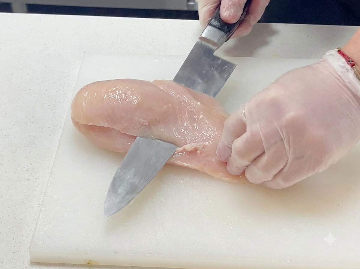 白いまな板の上で、手袋をした手が包丁を使って生肉の鶏むね肉を水平に慎重にスライスしているクローズアップ画像。