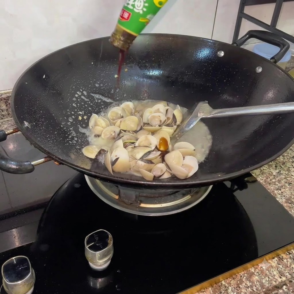 口が開いたばかりの白ハマグリと煮汁が入った中華鍋に、ボトルからオイスターソースを注いでいる様子