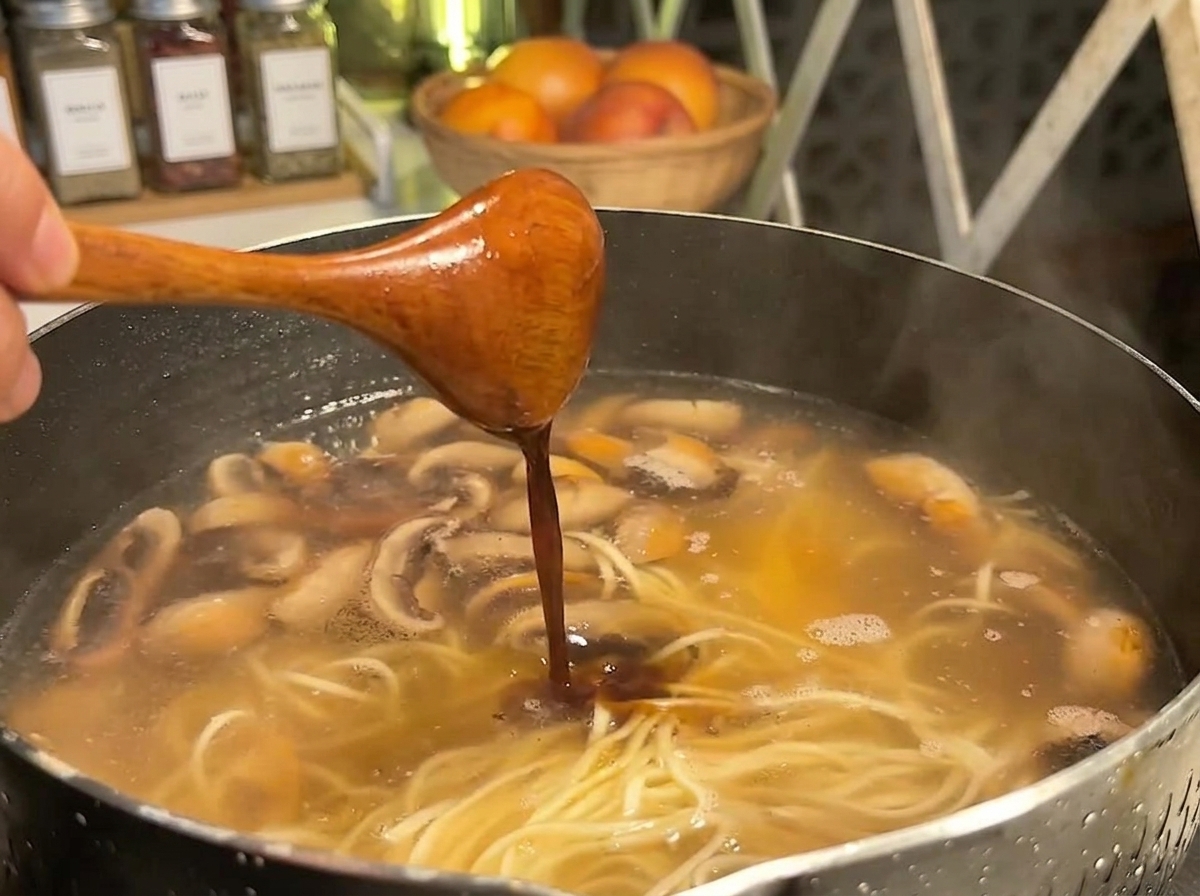 麺、スープ、スライスしたキノコが煮立っている鍋の中に、木製のスプーンから濃い調味料が注がれている様子。