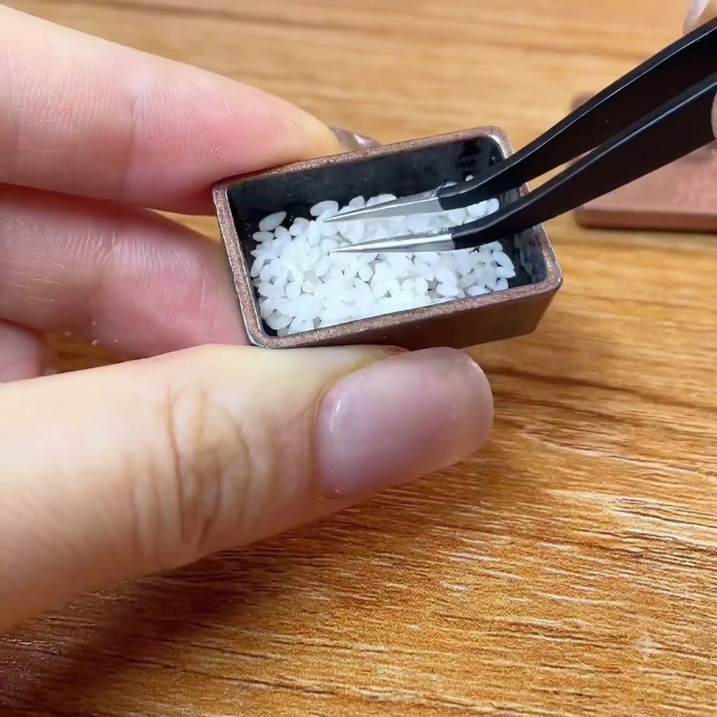 ピンセットを使って、小さな茶色の容器に極小の白い米粒を配置しているところ。